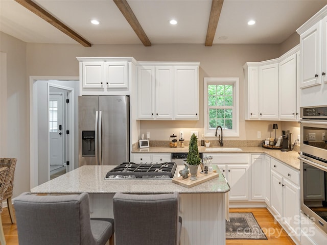 kitchen featuring stainless steel appliances, beamed ceiling, a sink, light wood-type flooring, and a kitchen island