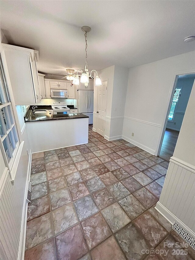 kitchen featuring white appliances, dark countertops, stone finish floors, pendant lighting, and wainscoting