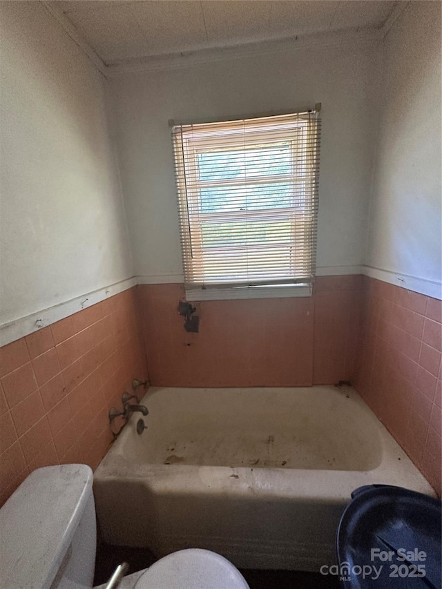 bathroom with toilet, ornamental molding, a garden tub, a wainscoted wall, and tile walls