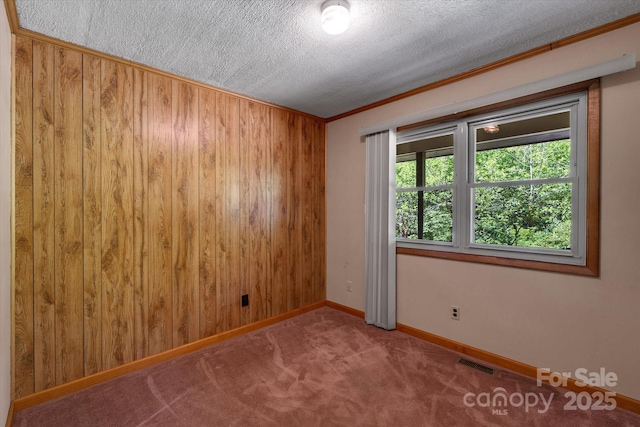 empty room featuring a textured ceiling, crown molding, carpet, baseboards, and wooden walls