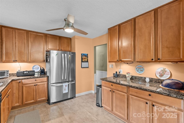 kitchen with freestanding refrigerator, a ceiling fan, dark stone countertops, brown cabinets, and a textured ceiling