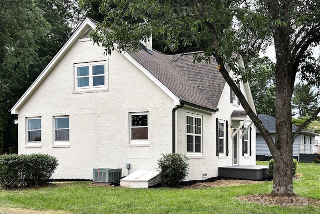 view of side of property featuring cooling unit, stucco siding, roof with shingles, and a yard