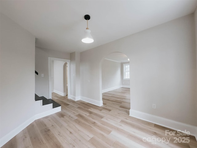 empty room featuring arched walkways, baseboards, light wood-type flooring, and stairs