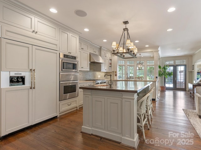 2410 Vernon Dr, Charlotte, NC 28211 - kitchen featuring built in appliances, under cabinet range hood, a kitchen island with sink, a chandelier, and dark wood-style flooring