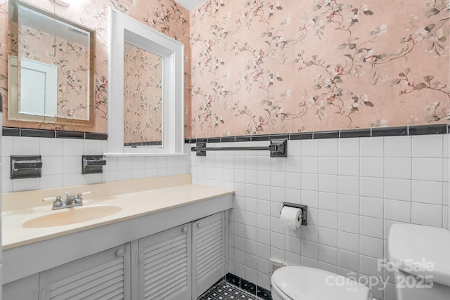 bathroom with wallpapered walls, toilet, a wainscoted wall, tile walls, and vanity