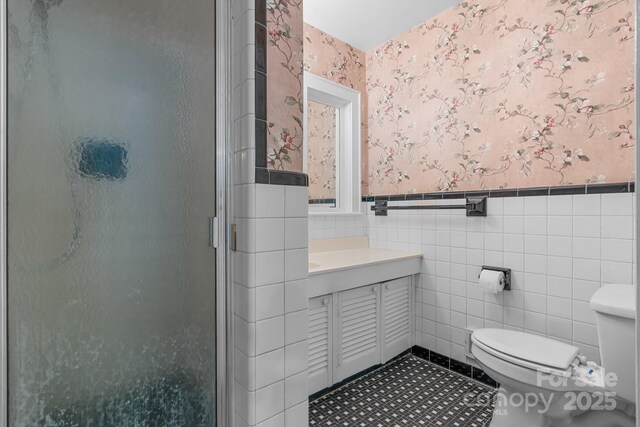 bathroom featuring wallpapered walls, toilet, a shower stall, tile walls, and a wainscoted wall