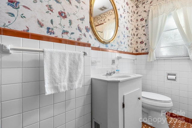 half bath with wallpapered walls, tile walls, toilet, wainscoting, and vanity
