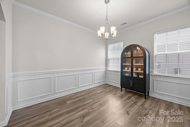 unfurnished dining area with a chandelier, ornamental molding, wood finished floors, wainscoting, and a decorative wall