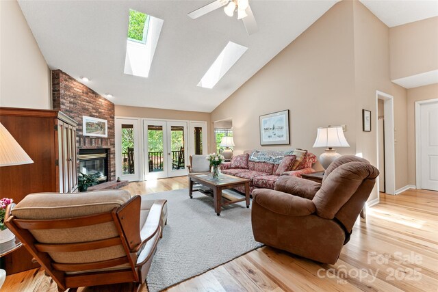 living area featuring high vaulted ceiling, a brick fireplace, light wood finished floors, a skylight, and ceiling fan