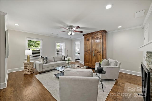 living area featuring a stone fireplace, ceiling fan, baseboards, crown molding, and dark wood-type flooring