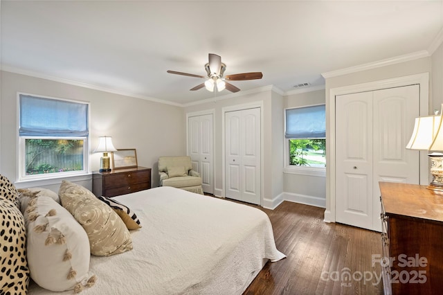 bedroom with two closets, dark wood-style floors, baseboards, ornamental molding, and a ceiling fan