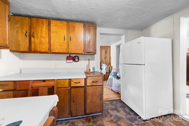 kitchen featuring freestanding refrigerator, brown cabinetry, light countertops, stone finish flooring, and a textured ceiling