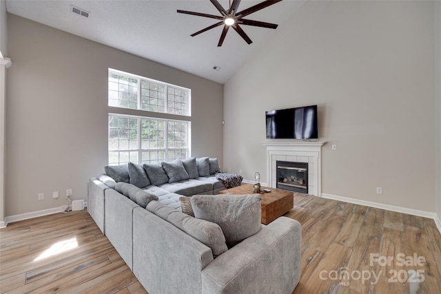 living area with high vaulted ceiling, ceiling fan, a tiled fireplace, light wood-style flooring, and baseboards