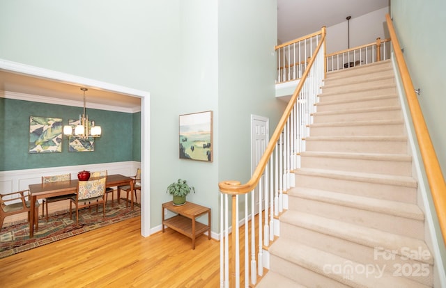 staircase with wood finished floors, a chandelier, a high ceiling, wainscoting, and crown molding