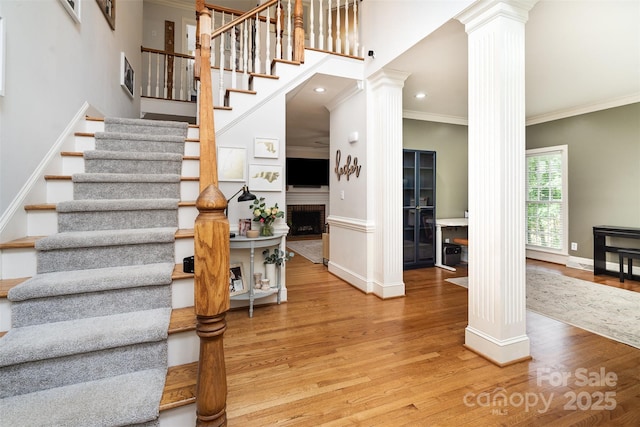 stairway featuring ornate columns, a brick fireplace, wood finished floors, crown molding, and baseboards