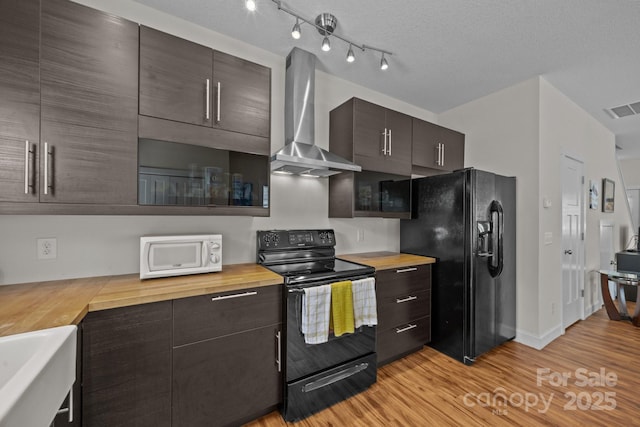 kitchen featuring black appliances, wall chimney exhaust hood, wooden counters, light wood finished floors, and dark brown cabinetry