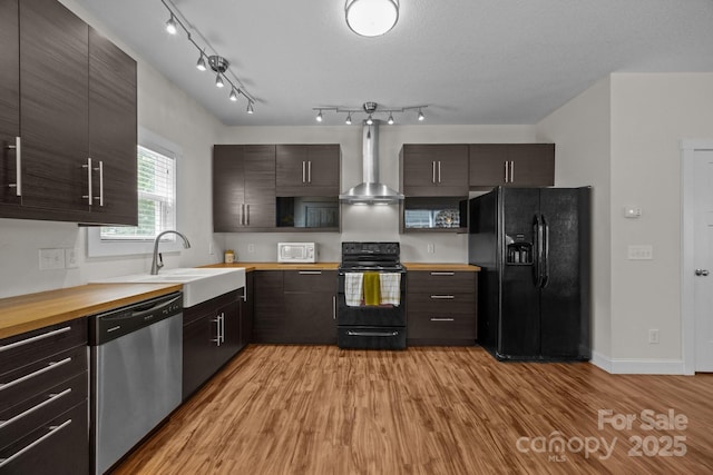 kitchen with black appliances, wall chimney range hood, a sink, light wood finished floors, and dark brown cabinets