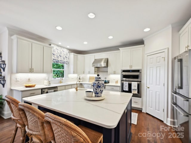 18419 Dembridge Dr, Davidson, NC 28036 - kitchen with wall chimney range hood, appliances with stainless steel finishes, a center island with sink, a kitchen bar, and dark wood finished floors