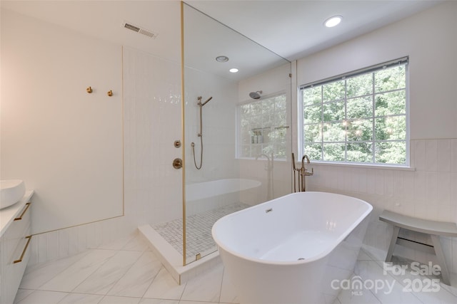 bathroom featuring a walk in shower, a soaking tub, vanity, wainscoting, and tile walls