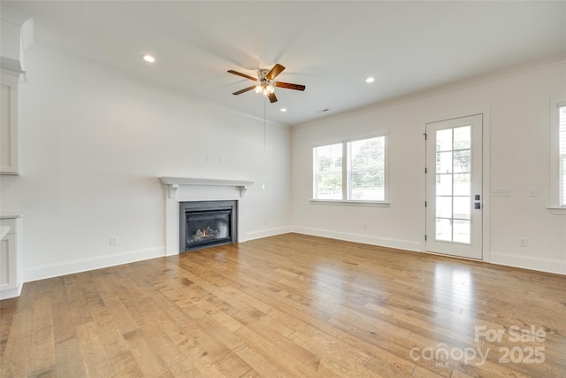 unfurnished living room with a warm lit fireplace, a ceiling fan, light wood finished floors, ornamental molding, and baseboards