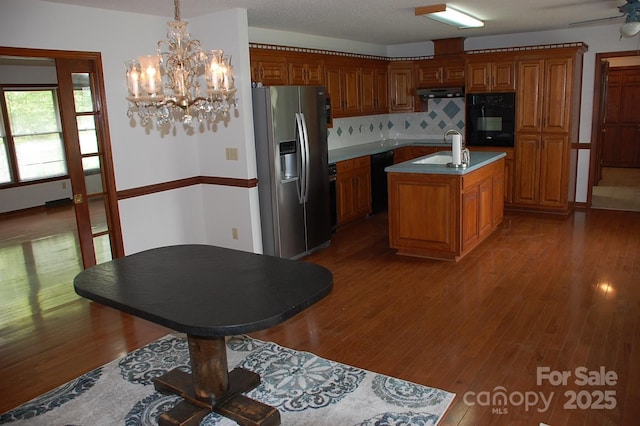 kitchen featuring black appliances, a center island with sink, brown cabinetry, tasteful backsplash, and under cabinet range hood