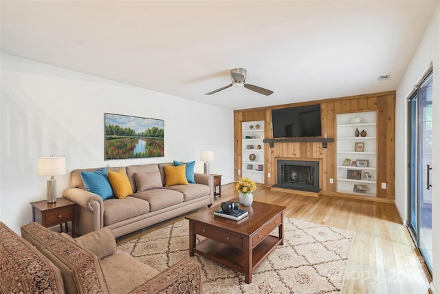 living room featuring light wood-style flooring, a ceiling fan, a fireplace with raised hearth, and built in features
