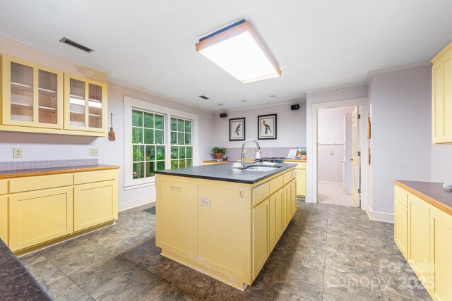 kitchen featuring a sink, cream cabinets, crown molding, dark countertops, and a center island with sink