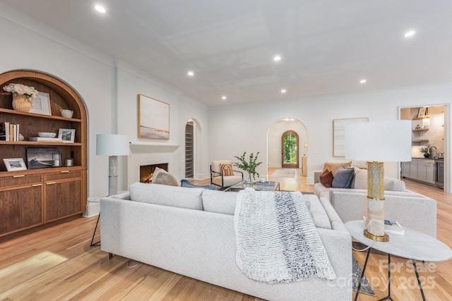 living room featuring arched walkways, crown molding, recessed lighting, light wood-style flooring, and a warm lit fireplace