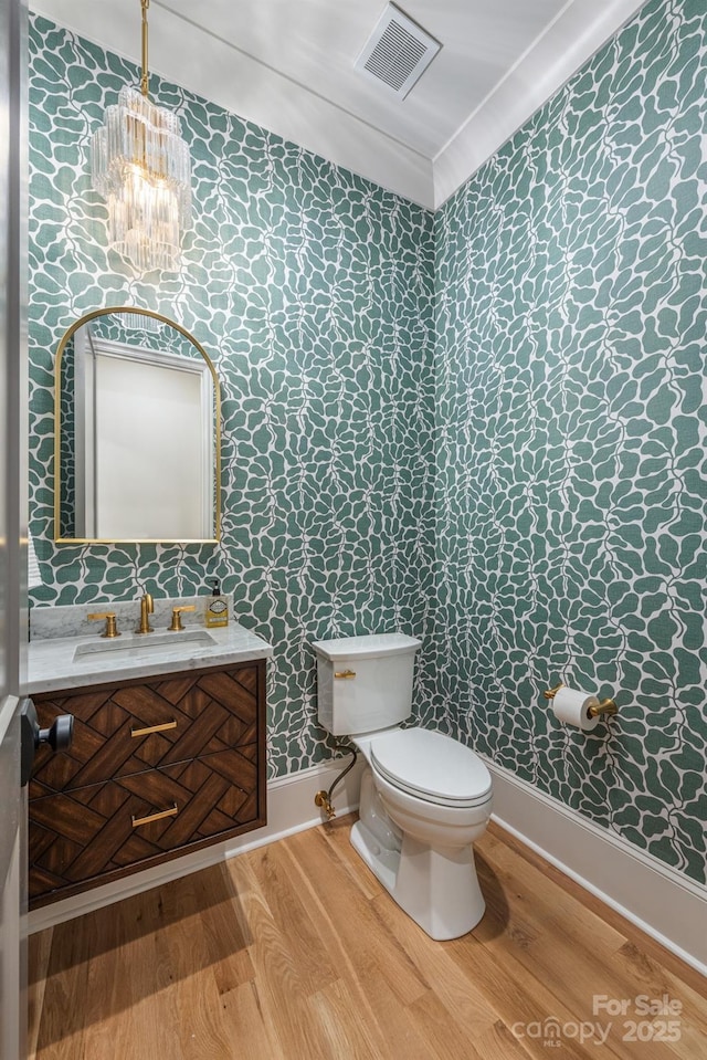 half bathroom with wallpapered walls, wood finished floors, a chandelier, toilet, and vanity