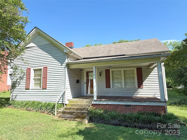 view of front facade with a chimney, covered porch, roof with shingles, and a front yard