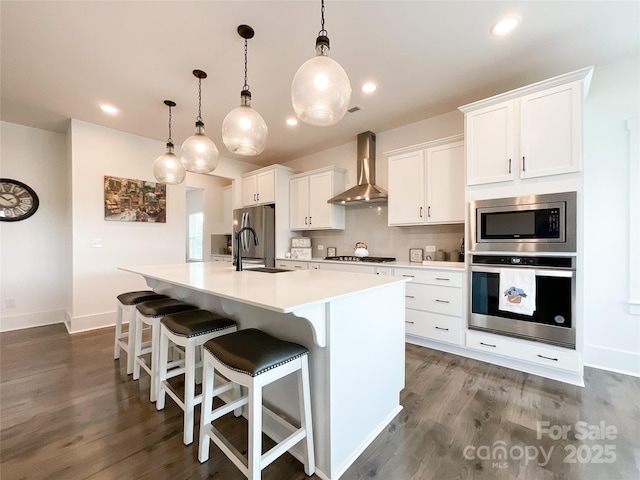 kitchen with appliances with stainless steel finishes, wall chimney exhaust hood, a center island with sink, dark wood-type flooring, and recessed lighting