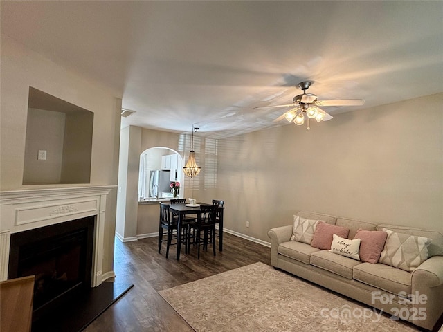 living area featuring arched walkways, a ceiling fan, dark wood-type flooring, a fireplace with raised hearth, and baseboards