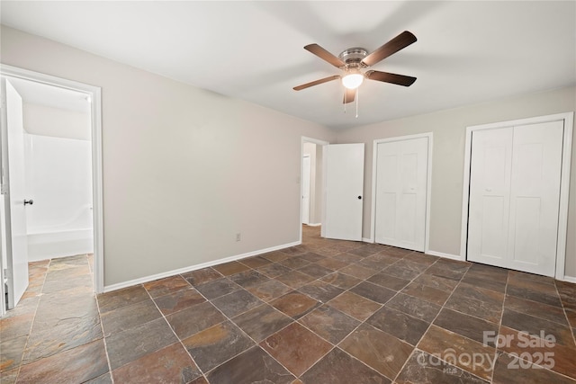 unfurnished bedroom with dark stone finish flooring, baseboards, a ceiling fan, and multiple closets