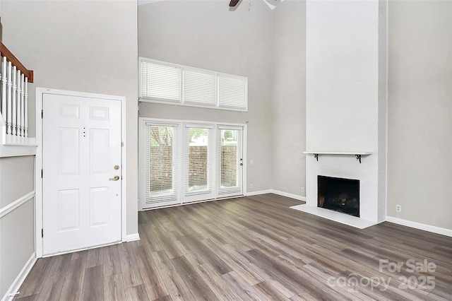 unfurnished living room with a towering ceiling, wood finished floors, a fireplace with flush hearth, baseboards, and a ceiling fan