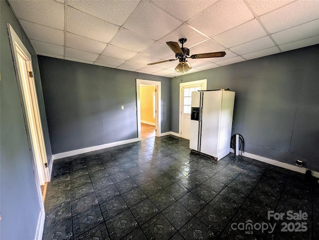 unfurnished bedroom with white refrigerator with ice dispenser, a drop ceiling, baseboards, and a ceiling fan