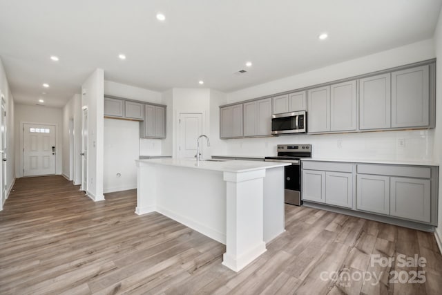 kitchen featuring stainless steel appliances, gray cabinetry, a center island with sink, light wood-type flooring, and recessed lighting