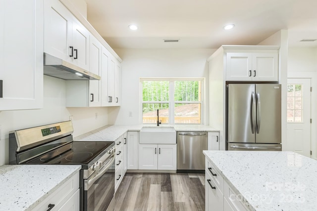 kitchen with appliances with stainless steel finishes, a sink, under cabinet range hood, wood finished floors, and white cabinetry