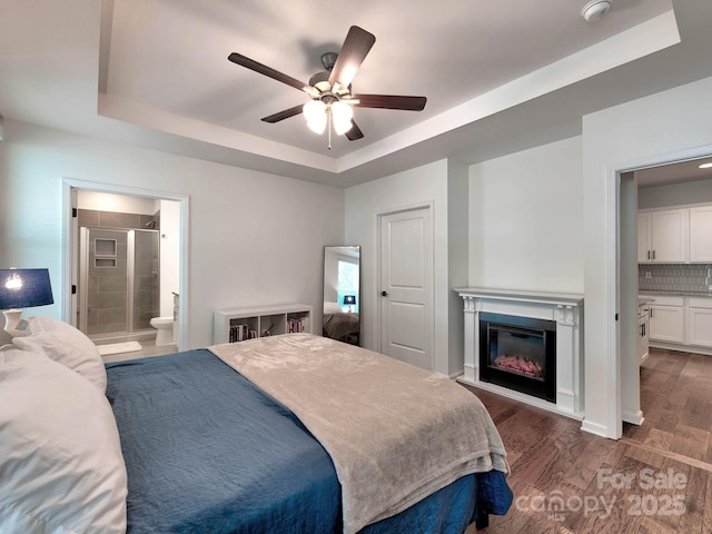 bedroom with a tray ceiling, dark wood-style flooring, a glass covered fireplace, connected bathroom, and a ceiling fan