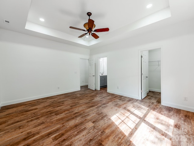 unfurnished bedroom featuring a tray ceiling, wood finished floors, baseboards, recessed lighting, and a walk in closet