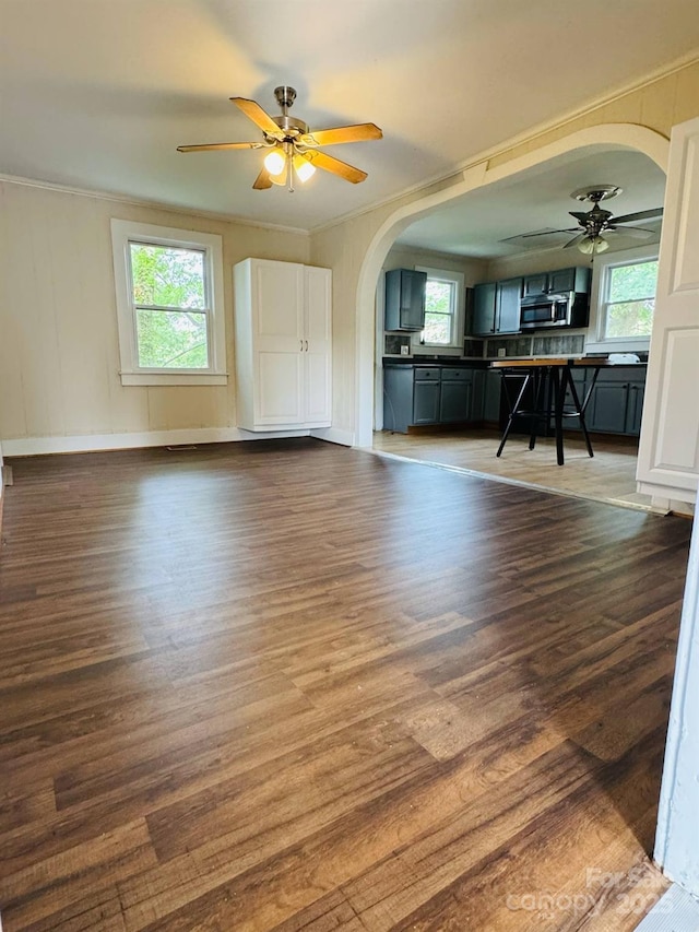 unfurnished living room featuring arched walkways, a ceiling fan, dark wood-type flooring, crown molding, and baseboards