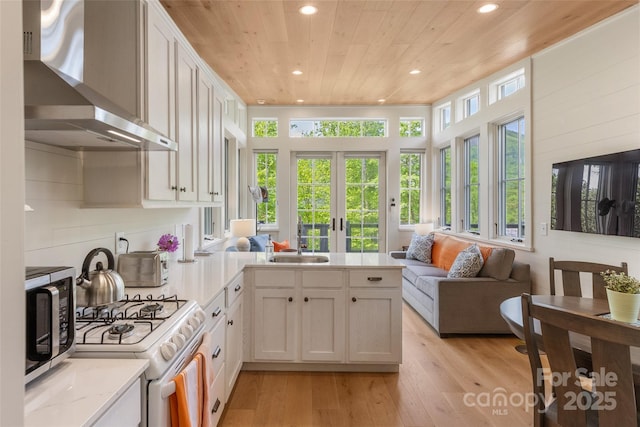 kitchen featuring wall chimney range hood, a peninsula, wooden ceiling, stainless steel microwave, and white range with gas stovetop