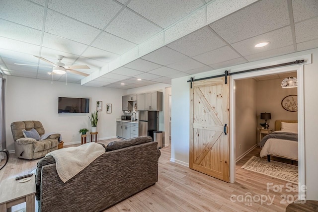 living room featuring a barn door, a drop ceiling, baseboards, light wood-style floors, and ceiling fan