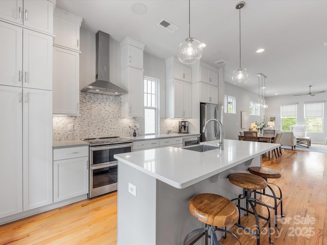 kitchen with wall chimney exhaust hood, light wood-style floors, appliances with stainless steel finishes, a sink, and an island with sink