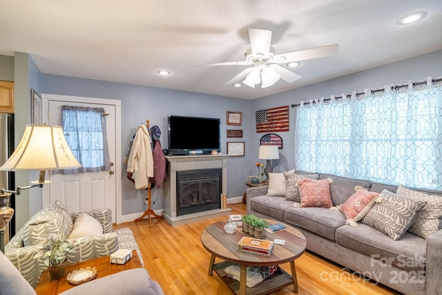 living area featuring light wood-type flooring, ceiling fan, a glass covered fireplace, baseboards, and recessed lighting