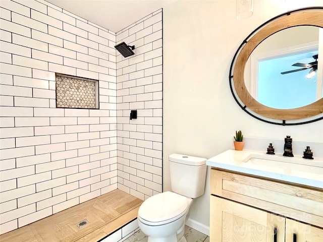 bathroom with toilet, tiled shower, vanity, ceiling fan, and baseboards
