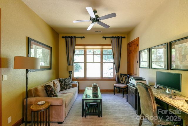 living room with a desk, light carpet, a ceiling fan, a textured wall, and baseboards