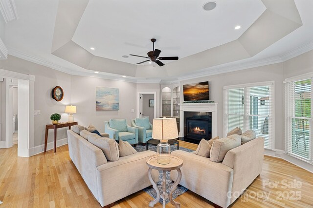 17308 Players Ridge Dr, Cornelius, NC 28031 - living room featuring a raised ceiling, light wood-type flooring, a glass covered fireplace, a ceiling fan, and baseboards
