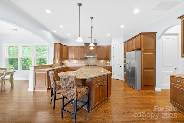 2289 Pine Valley Dr, Denver, NC 28037 - kitchen with stainless steel appliances, under cabinet range hood, a center island, brown cabinets, and dark wood-style flooring