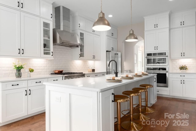 3009 Ocaso Ct, Matthews, NC 28104 - kitchen featuring wall chimney exhaust hood, backsplash, a kitchen island with sink, arched walkways, and light wood-style flooring