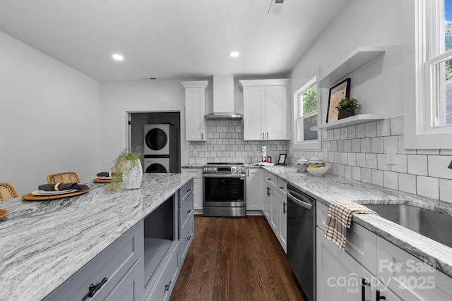 1922 Thomas Ave, Charlotte, NC 28205 - kitchen featuring wall chimney exhaust hood, stacked washing machine and dryer, appliances with stainless steel finishes, light stone counters, and dark wood-style flooring